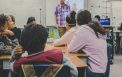 Students seated at desks in a diverse classroom, listening to a teacher standing at the front near a whiteboard.