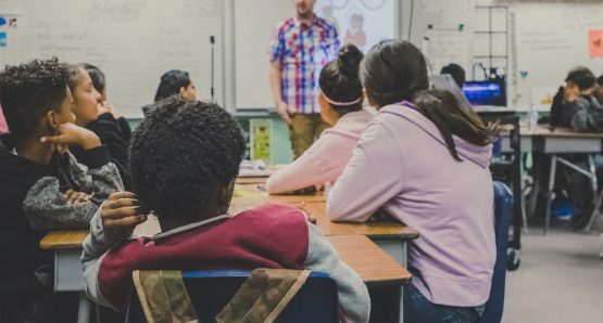 Students seated at desks in a diverse classroom, listening to a teacher standing at the front near a whiteboard.